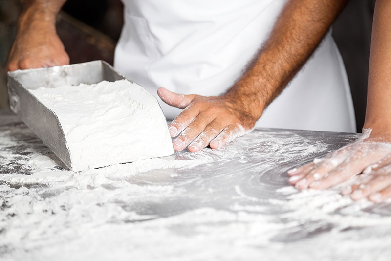 Metal commercial kitchen counter with person scooping large amount of flour onto surface